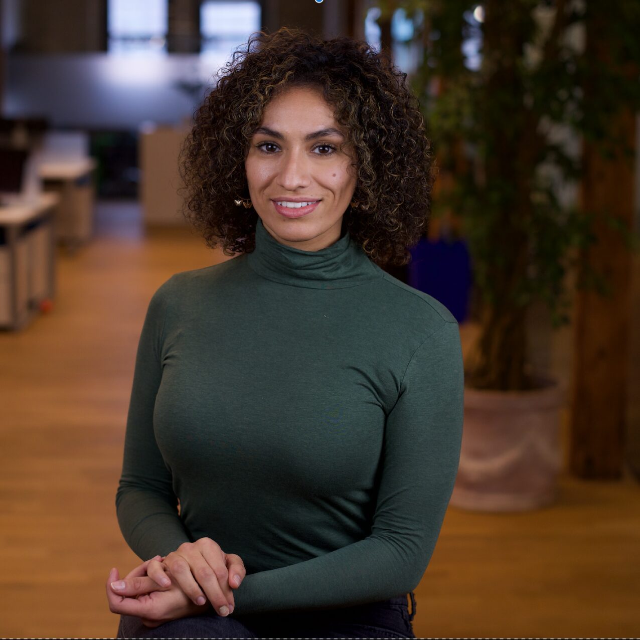 A woman with curly hair wearing a green turtleneck sweater in a cozy indoor setting.