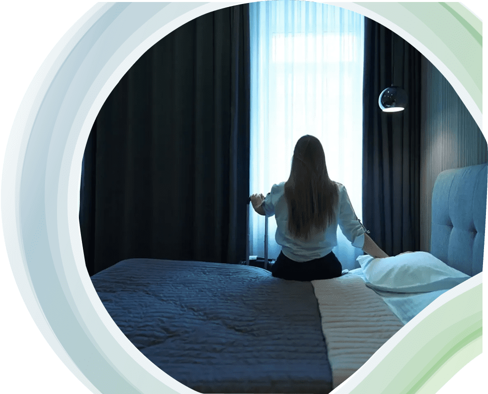 Woman sitting on bed opening curtains to morning light.