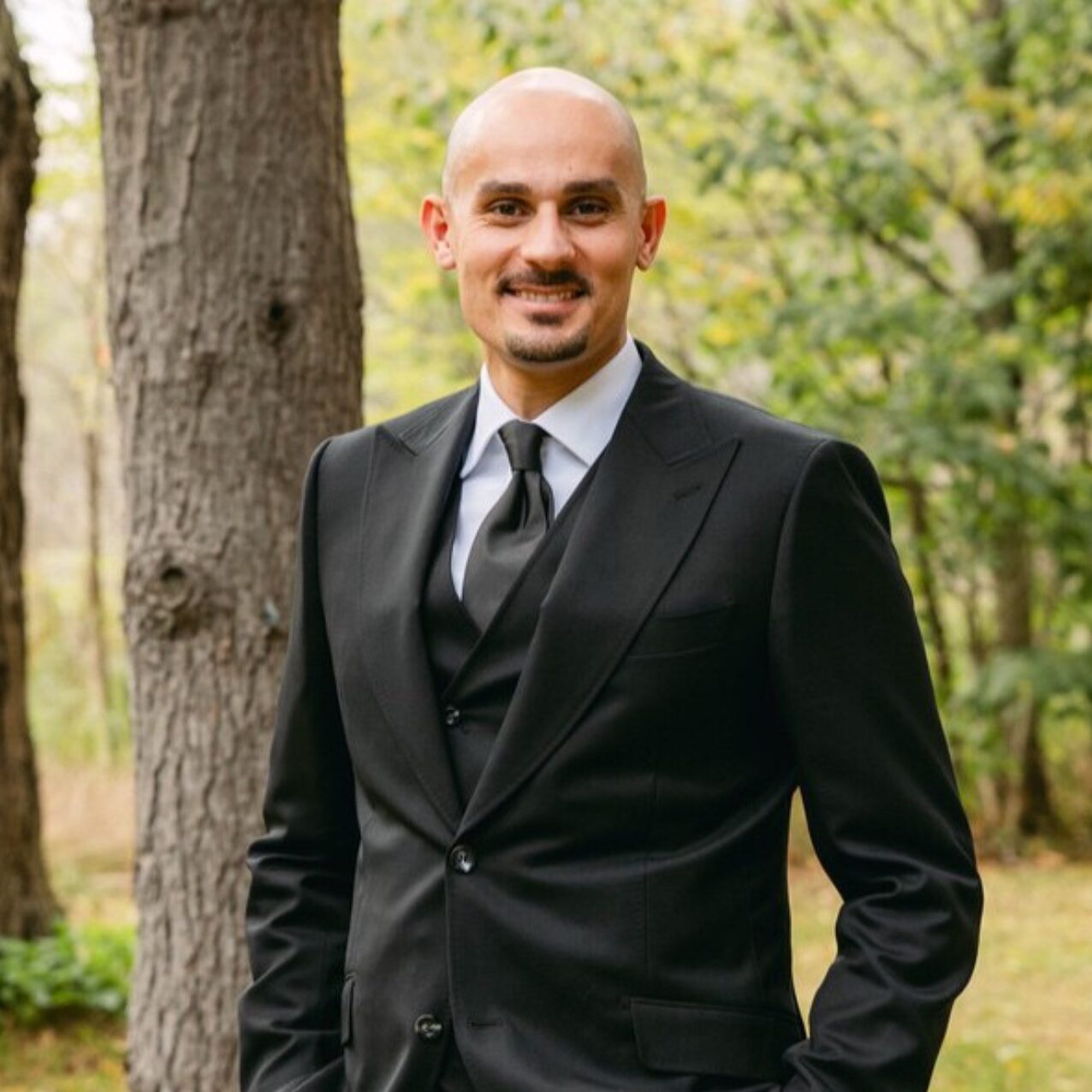 Man in a black suit standing outdoors near trees.